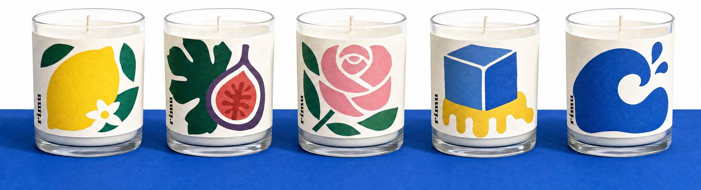 Five clear glass candle jars on a cobalt shelf, each with a cut-paper label: a lemon, a fig leaf, a pink rose, a blue sugar cube and a wave.